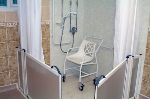 Salle de bain comprenant une douche PMR équipée de rampes, fauteuil et douchette parfaitement adaptée pour une personne à mobilité réduite